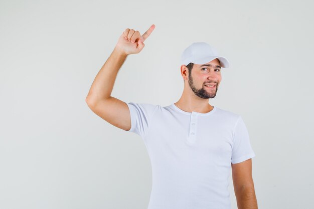 Молодой человек указывая вверх, улыбаясь в футболке, кепке и выглядя довольным, вид спереди.