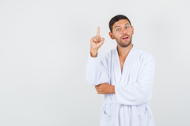 Young man pointing up in white bathrobe and looking smart , front view.