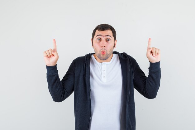 Young man pointing up with index fingers in white t-shirt and zip-front black hoodie and looking surprised. front view.