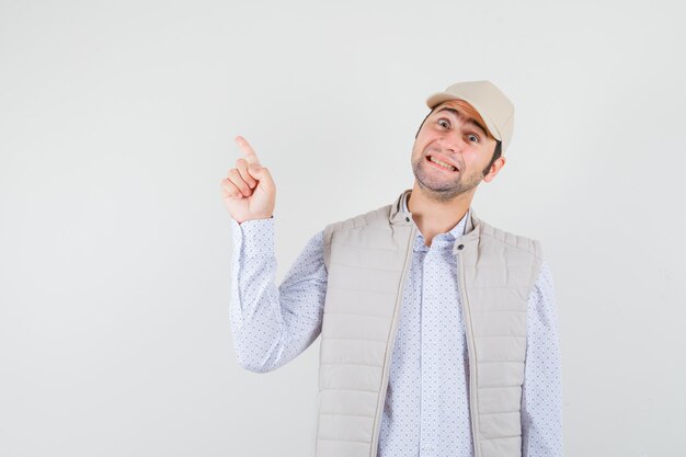 Young man pointing up with one hand in beige jacket and cap and looking happy , front view.