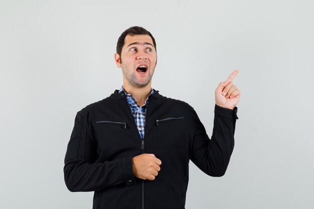 Young man pointing at upper right corner in shirt, jacket and looking hopeful , front view.