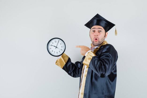 Young man pointing at wall clock in graduate uniform and looking amazed. .