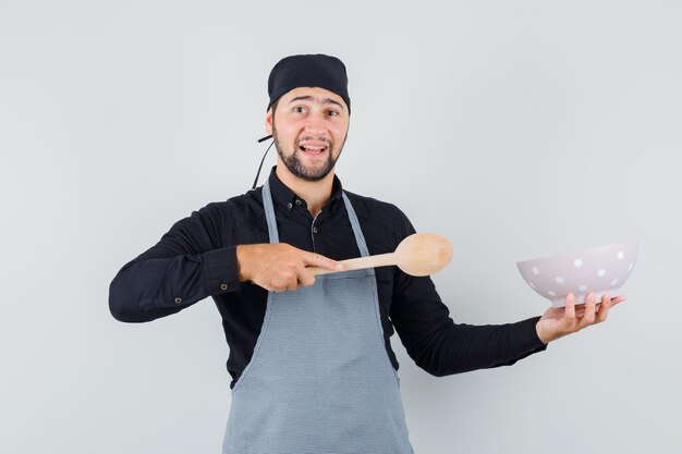 Young man pointing wooden spoon at bowl in shirt, apron and looking cheery. front view.