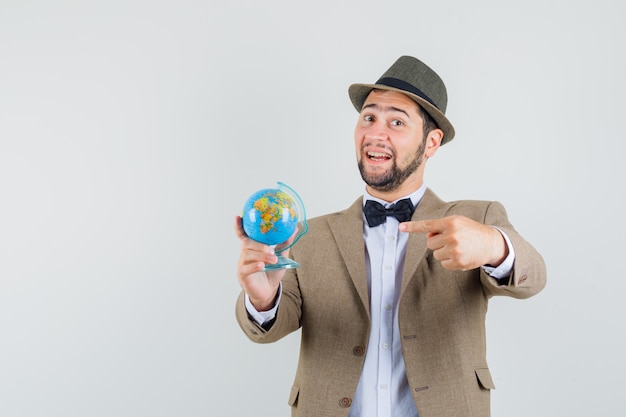 Free photo young man pointing at world globe in suit, hat and looking cheery. front view.