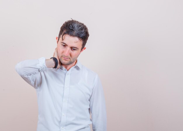 Young man posing while holding hand on neck in white shirt and looking pensive