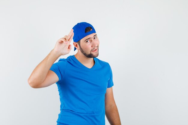 Young man posing while holding two fingers together in blue t-shirt and cap and looking funny