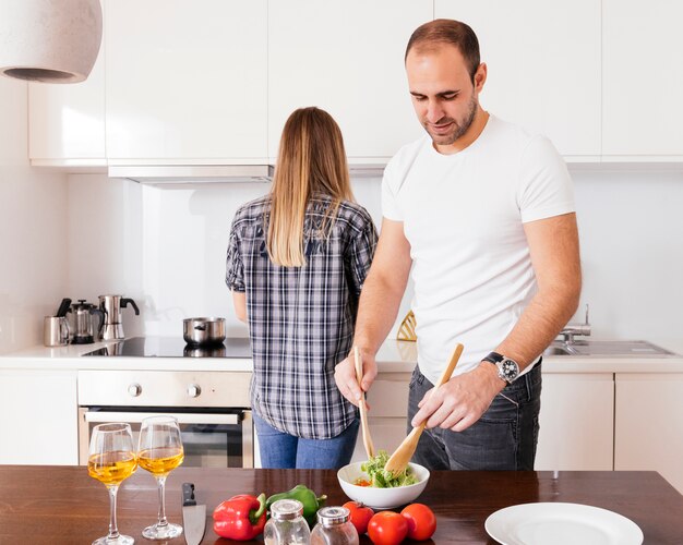 Young man preparing the salad and her wife standing behind him cooking food in the kitchen