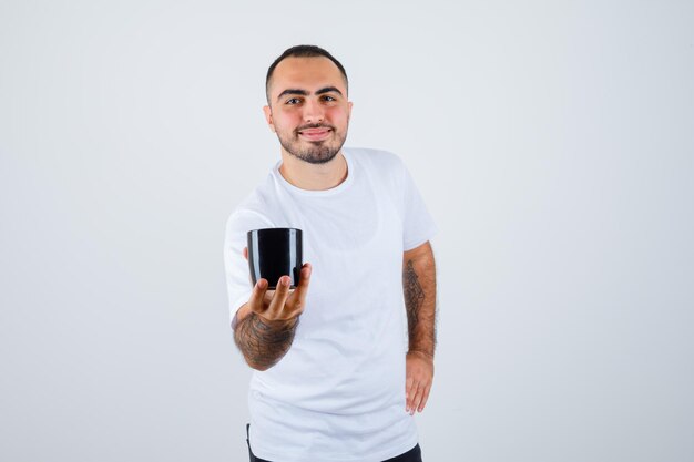 Young man presenting cup of tea while holding hand on waist in white t-shirt and black pants and looking happy