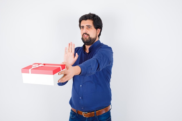 Young man presenting gift box and showing stop sign in blue shirt and jeans and looking scared , front view.