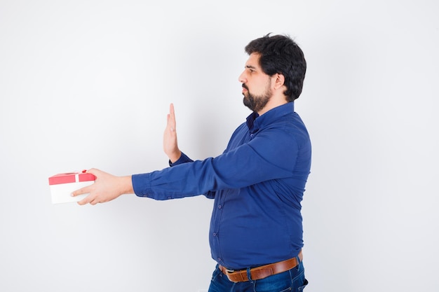 Young man presenting gift box and showing stop sign in blue shirt and jeans and looking scared , front view.