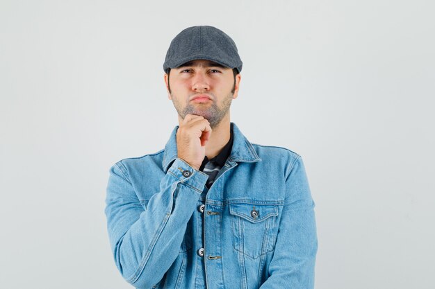 Young man propping chin on hand in cap, t-shirt, jacket and looking pensive. front view.