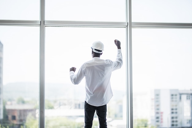 Молодой человек в защитном подоле стоит перед панорамными окнами с поднятыми руками победы и успеха