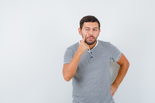 Young man pulling down his eyelid in t-shirt and looking displeased. front view.