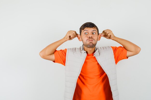 Young man pulling ears with fingers, blowing cheeks in t-shirt, jacket and looking funny