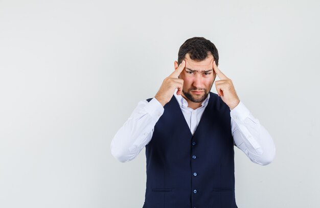 Young man pulling skin on his temples in shirt and vest and looking exhausted