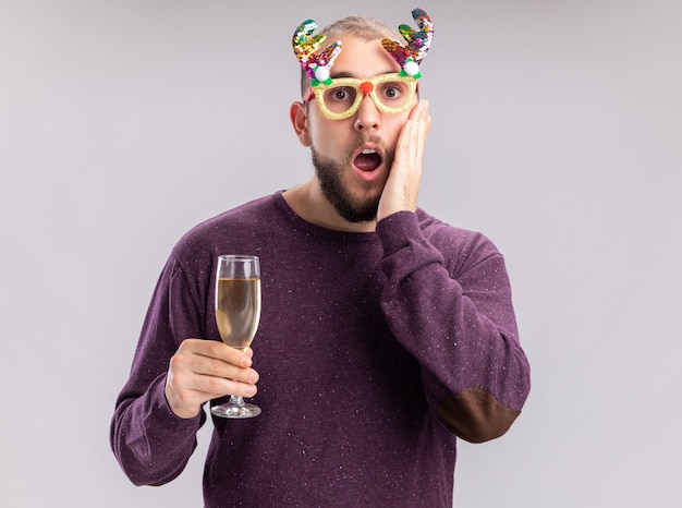 Молодой человек в фиолетовом свитере и забавных очках с бокалом шампанского, глядя в камеру, изумлен и удивлен, стоя на белом фоне