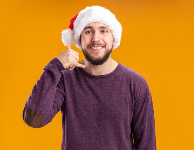 Молодой человек в фиолетовом свитере и шляпе санта-клауса, счастливый и позитивный, показывает жест "зови меня", стоя над оранжевой стеной