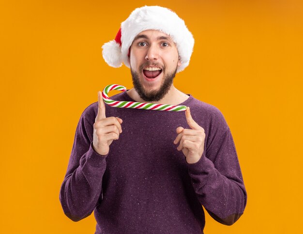 Young man in purple sweater and santa hat holding candy cane looking at camera with happy face standing over orange background