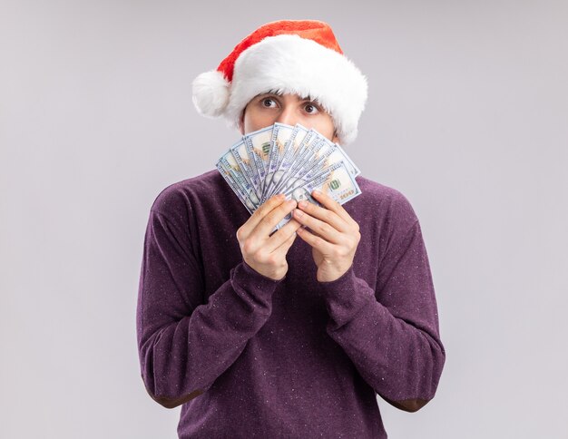 Young man in purple sweater and santa hat holding cash covering face with money looking aside worried standing over white background