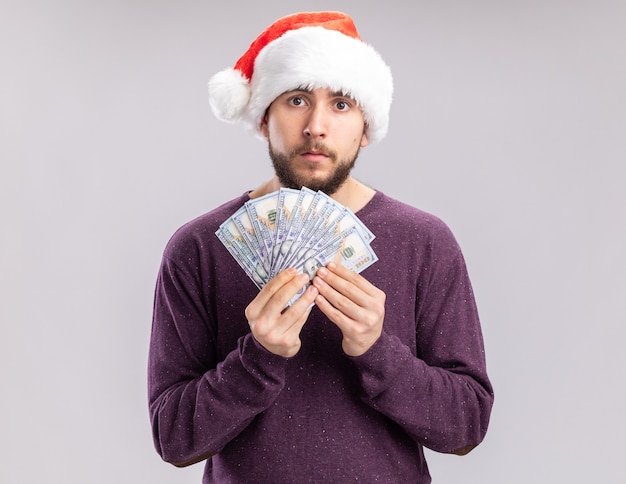 보라색 스웨터와 흰색 배경 위에 서 심각한 얼굴로 카메라를 찾고 현금을 들고 산타 모자에 젊은 남자