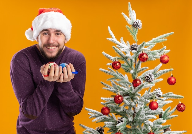 Giovane uomo in maglione viola e cappello santa tenendo le palle di natale sorridente felice ed emozionato in piedi accanto all'albero di natale su sfondo arancione