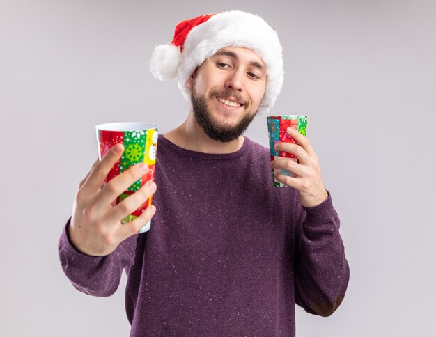 Young man in purple sweater and santa hat holding colorful paper cups looking at them smiling cheerfully standing over white background