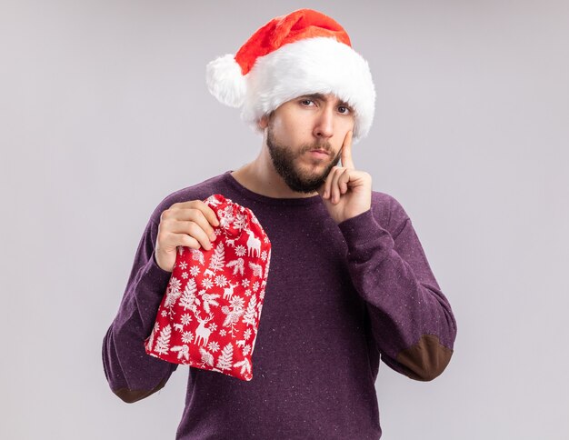 보라색 스웨터와 산타 모자 흰색 배경 위에 서 회의적인 표정으로 카메라를 찾고 선물 빨간 가방을 들고 젊은 남자