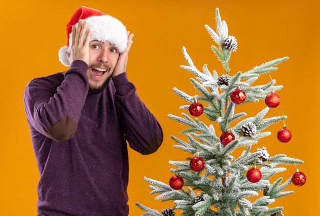 Young man in purple sweater and santa hat looking amazed and surprised standing next to christmas tree over orange background