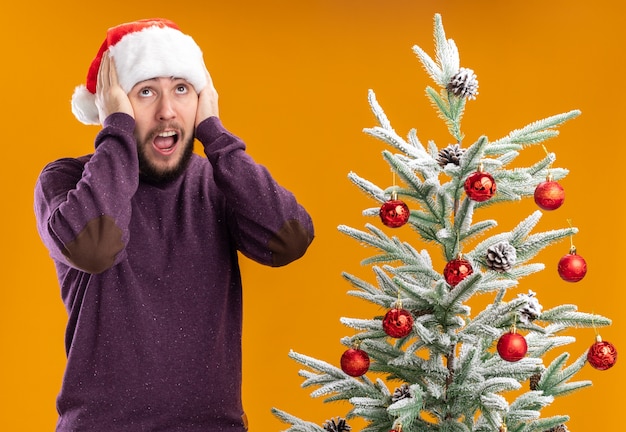 Молодой человек в фиолетовом свитере и новогодней шапке выглядит изумленным и удивленным, стоя рядом с елкой на оранжевом фоне