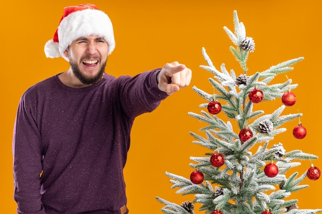 オレンジ色の背景の上のクリスマスツリーの横に立っている何かを人差し指で指しているイライラした表情で脇を見て紫色のセーターとサンタ帽子の若い男