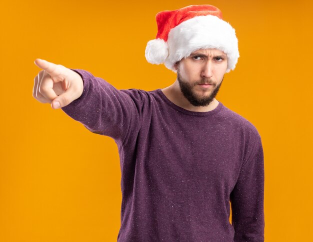 Young man in purple sweater and santa hat looking aside with serious face pointing with index finger at something standing over orange background