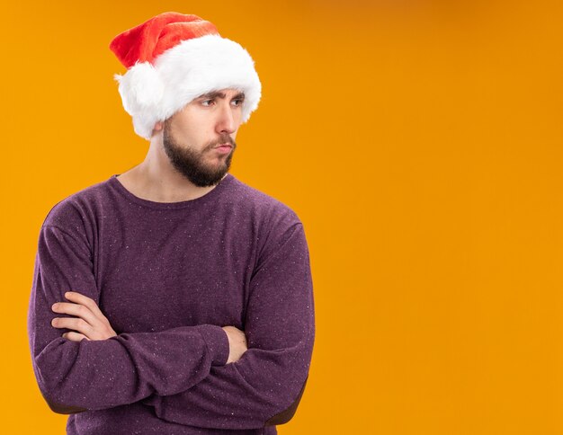 オレンジ色の背景の上に立っている深刻な顔で脇を見ている紫色のセーターとサンタ帽子の若い男