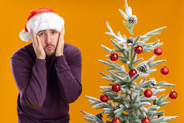 Young man in purple sweater and santa hat looking at camera confused and very anxious next to christmas tree over orange background