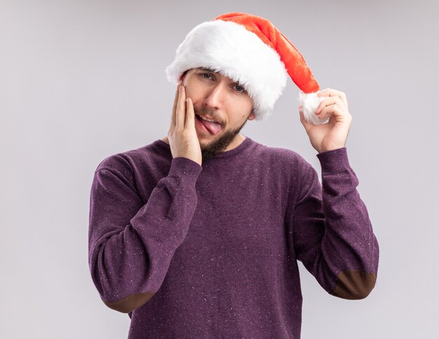 Young man in purple sweater and santa hat looking at camera happy and joyful sticking out tongue standing over white background