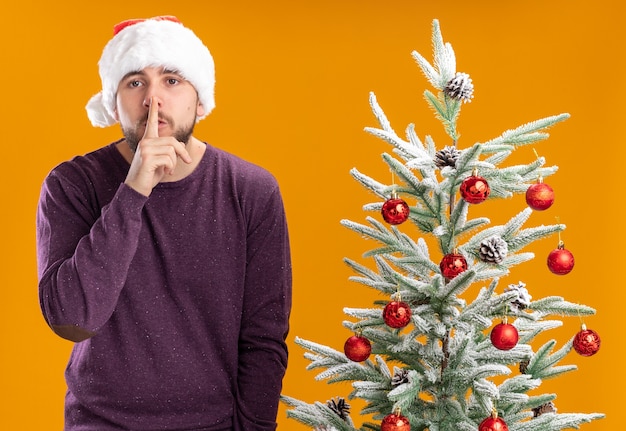オレンジ色の背景の上のクリスマスツリーの横に沈黙のジェスチャーを作るカメラを見て紫色のセーターとサンタ帽子の若い男