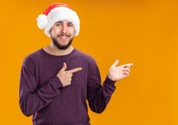 Young man in purple sweater and santa hat looking at camera smiling cheerfully pointing with index fingers to the side standing over orange background