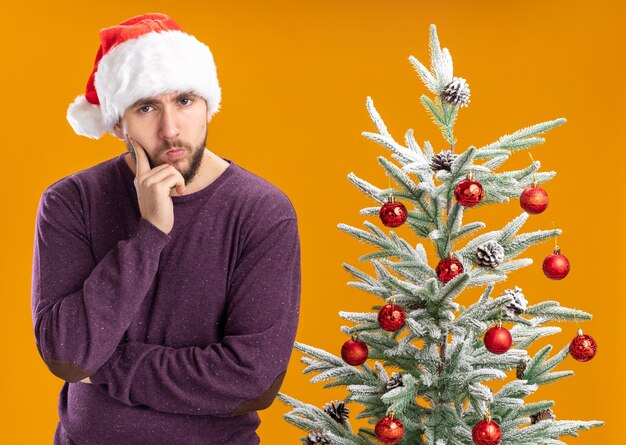 オレンジ色の背景の上のクリスマスツリーの横に物思いにふける表情でカメラを見て紫色のセーターとサンタ帽子の若い男