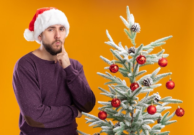 Young man in purple sweater and santa hat looking at camera with skeptic expression on face standing next to christmas tree over orange background