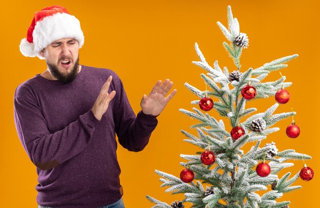 Young man in purple sweater and santa hat looking at christmas tree with disgusted expression making defense gesture standing over orange wall