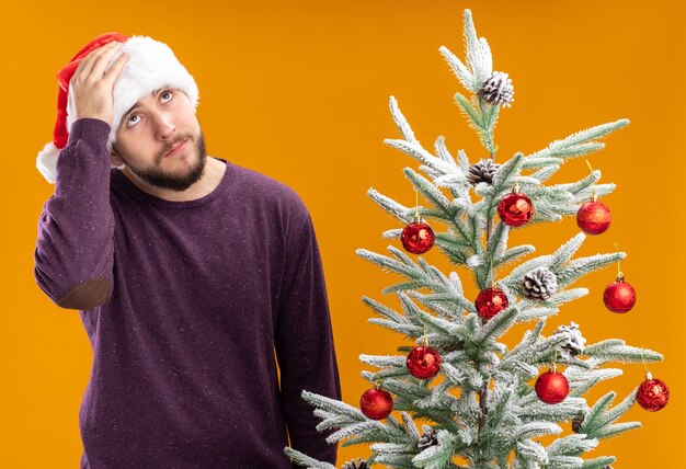 Young man in purple sweater and santa hat looking confused and very anxious standing next to christmas tree over orange background