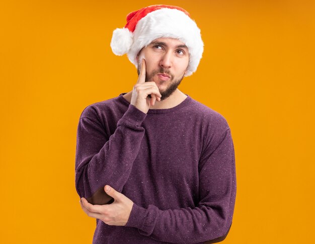 Молодой человек в фиолетовом свитере и шляпе санта-клауса смотрит вверх с пальцем на щеке с задумчивым выражением лица, думая, стоя на оранжевом фоне