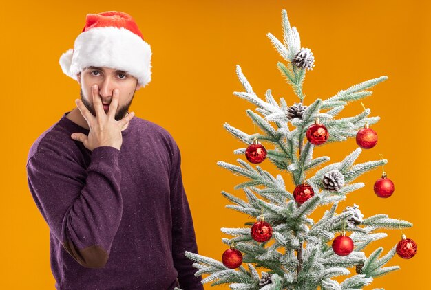 Young man in purple sweater and santa hat  pulling eyes down with fingers tired and bored standing next to christmas tree over orange wall