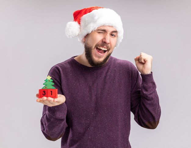 Young man in purple sweater and santa hat showing cubes with new year clenching fist happy and excited standing over white background