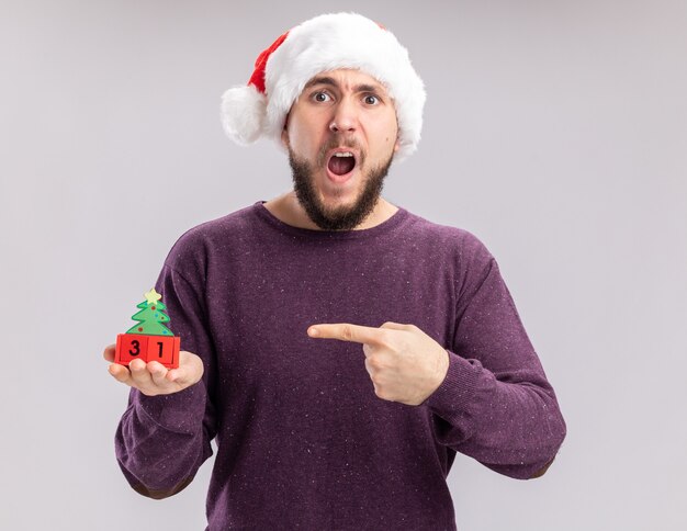 Молодой человек в фиолетовом свитере и новогодней шапке показывает кубики с новогодней датой, указывая на нее указательным пальцем, изумленный и удивленный, стоя на белом фоне