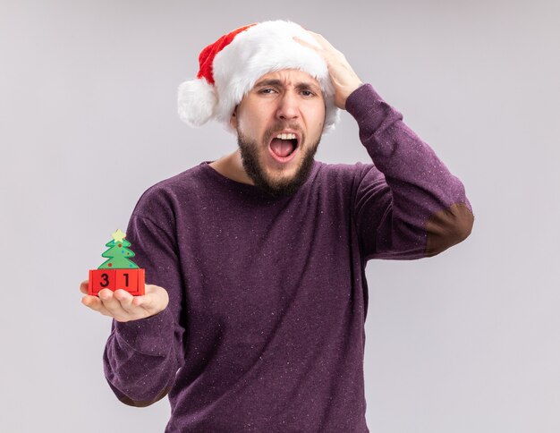 Молодой человек в фиолетовом свитере и шляпе санта-клауса показывает кубики с новогодней датой, кричит с разочарованным выражением лица, стоя на белом фоне
