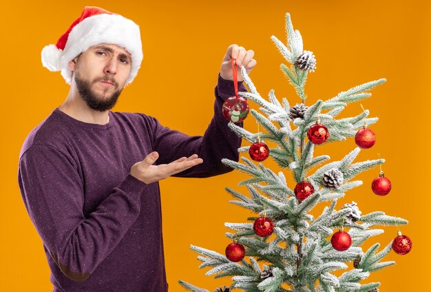 Young man in purple sweater and santa hat standing next to christmas tree holding toy hanging it on tree with serious face over orange background