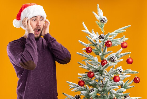 보라색 스웨터와 산타 모자 서있는 젊은 남자가 나무를보고 크리스마스 트리 옆에 서서 오렌지 배경 위에 놀랐고 놀랐습니다.