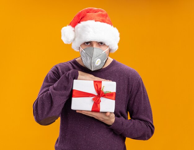 Young man in purple sweater and santa hat wearing facial protecrive mask holding present looking at camera with serious face standing over orange background