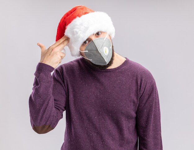 Free photo young man in purple sweater and santa hat wearing facial protective mask making pistol gesture with fingers over temple tired and bored standing over white background