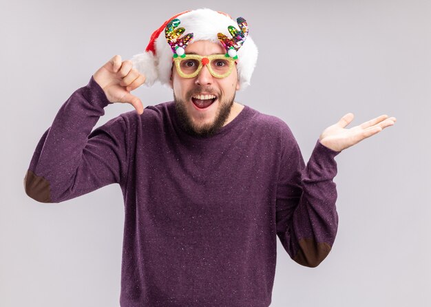 Young man in purple sweater and santa hat wearing funny glasses  happy and joyful smiling cheerfully showing thumbs down standing over white wall